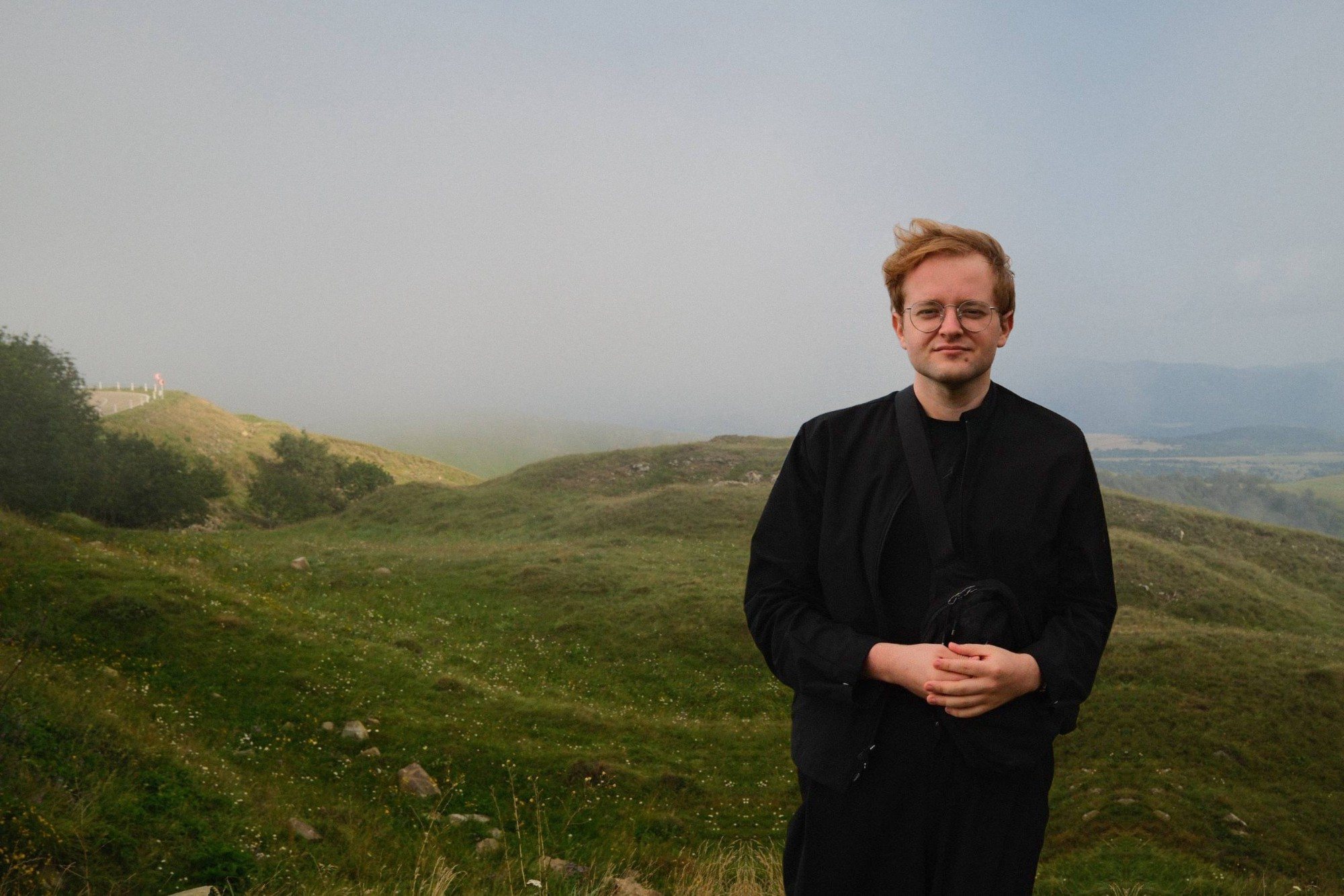 Pavel Rysych in a black jacket and round glasses, standing on a green hillside in mountain fog